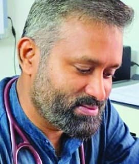Dr. Shavas MM, wearing a blue shirt and a stethoscope, at his desk