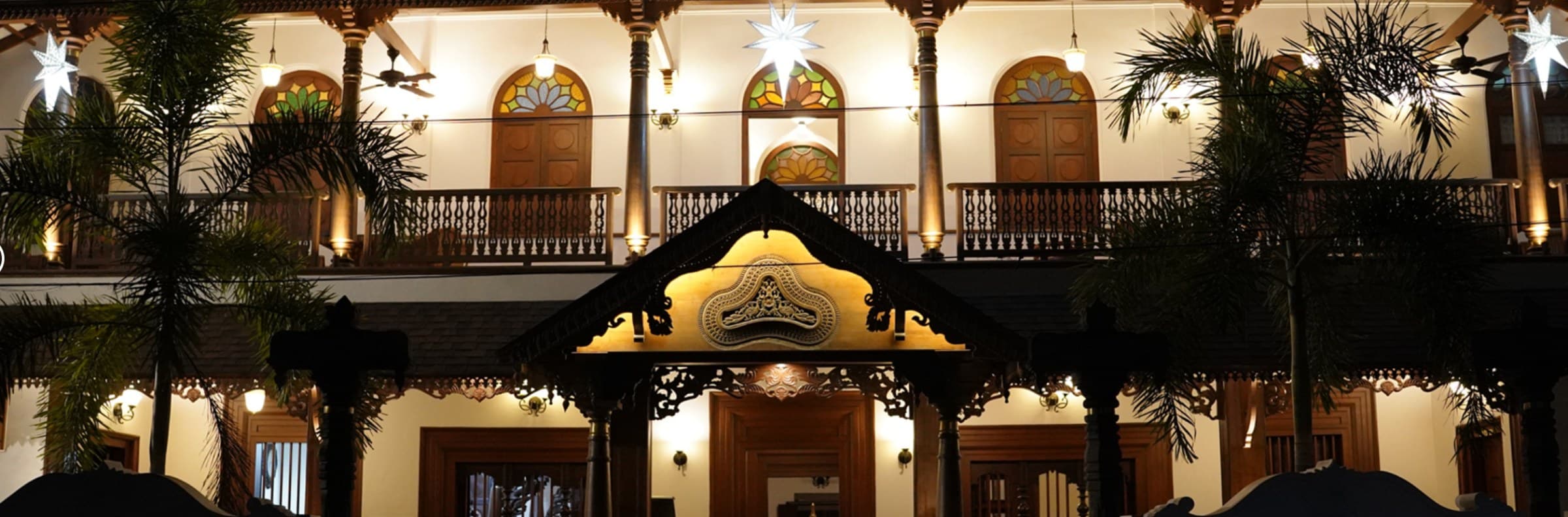 Villa De 1945 illuminated at night — a carved timber gable with a brass medallion above the entrance, stained-glass fanlights along the upper verandah, turned balusters and star lanterns
